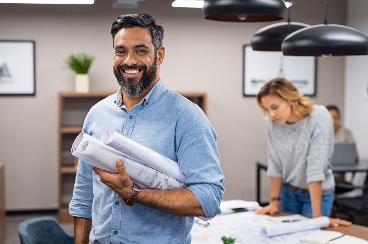 Smiling male architect or designer holding rolled blueprints in a modern office setting while a colleague works in the background