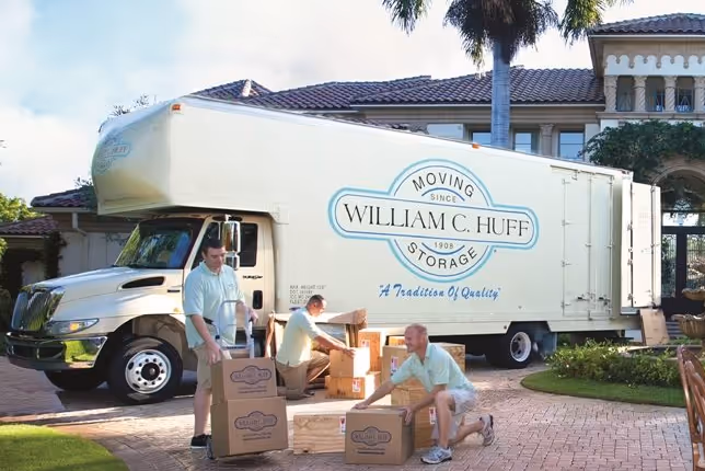 Three professional movers unloading boxes from a William C. Huff moving truck in front of a luxury home