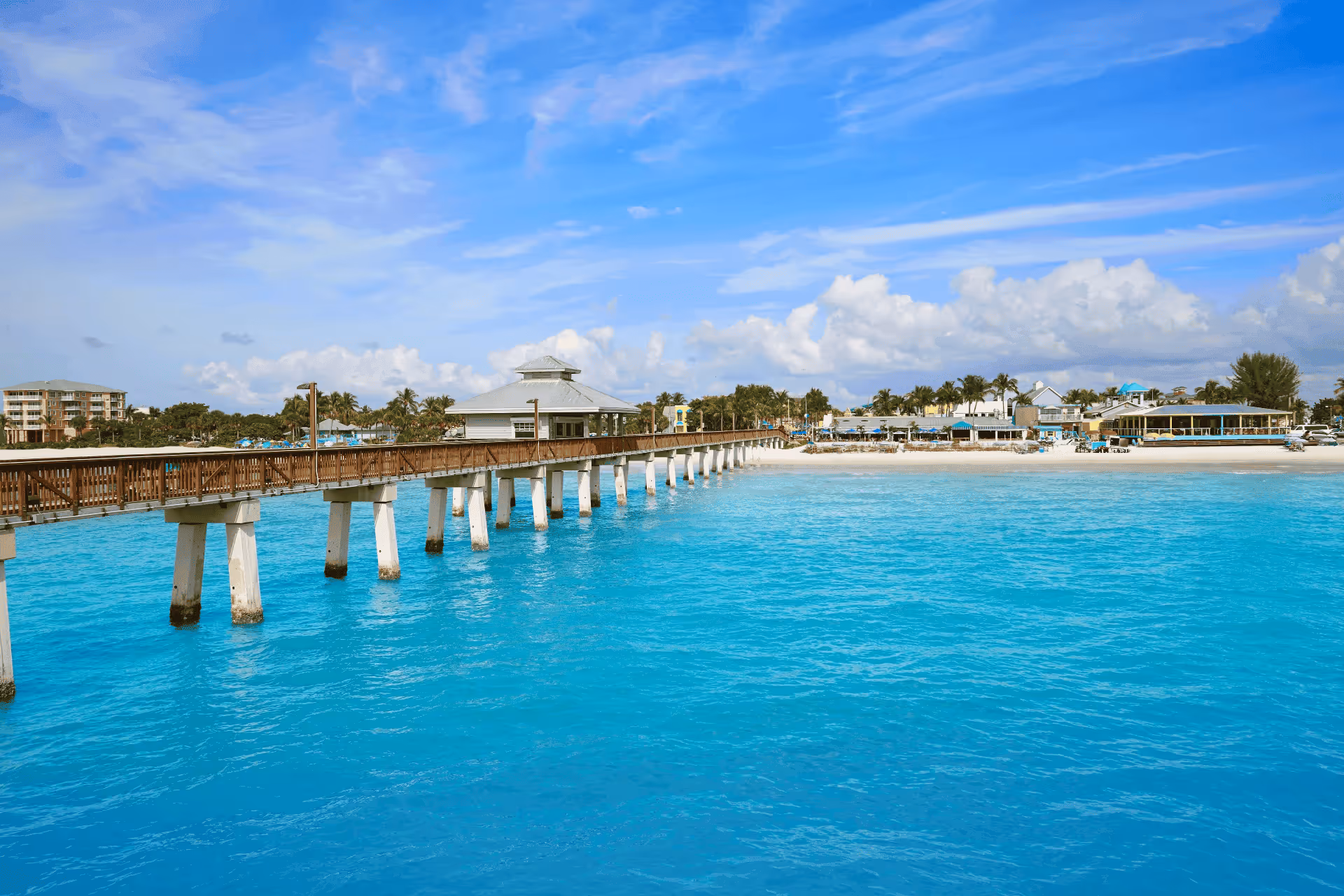 Long wooden pier extending over turquoise ocean water toward a sunny beach resort