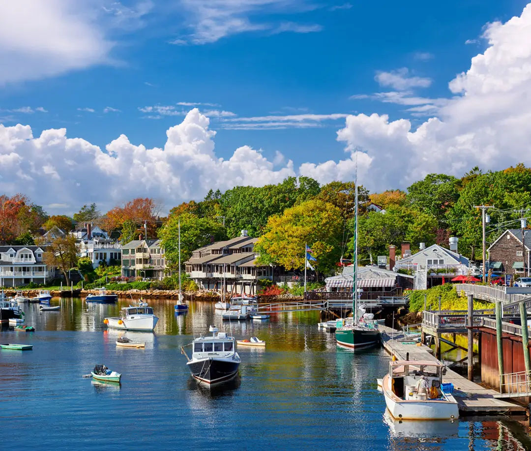 Boats anchored in the calm blue waters of Perkins Cove in Ogunquit, Maine, surrounded by lush green trees and coastal buildings under a bright, cloudy sky