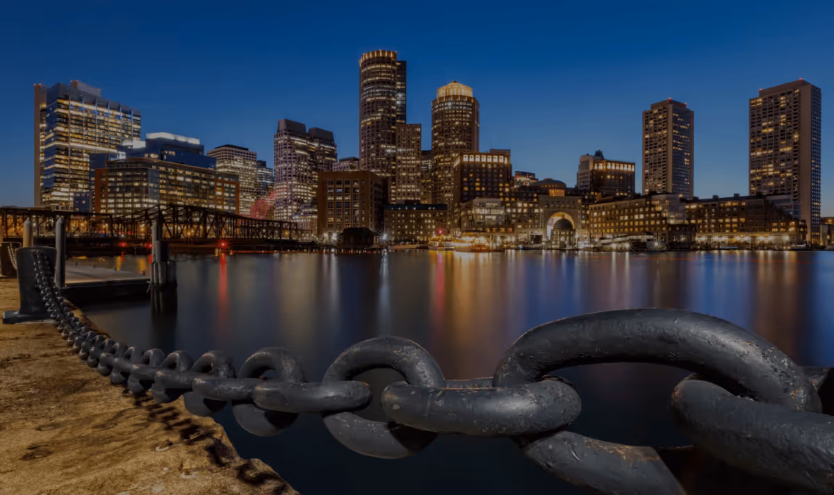 Boston skyline at dusk with harbor water reflections and a large foreground chain
