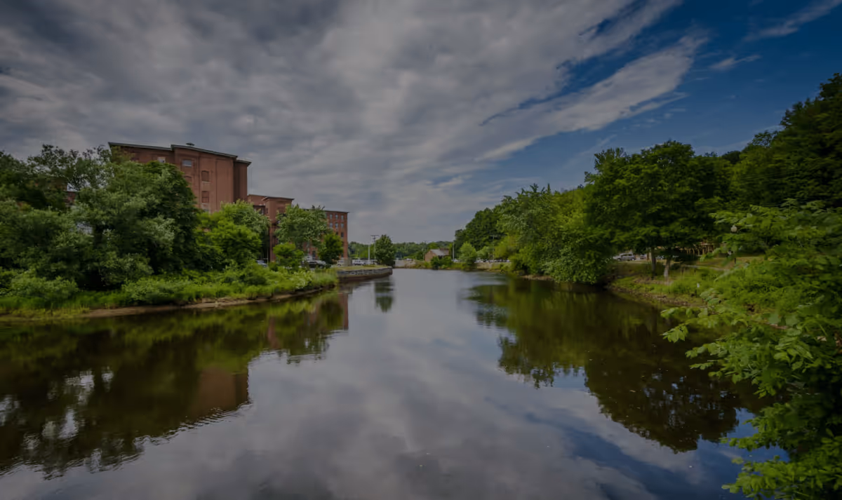 A scenic view of the Cocheco River flowing through Dover, New Hampshire, with historic brick mill buildings and lush green trees reflected in the water under a cloudy sky