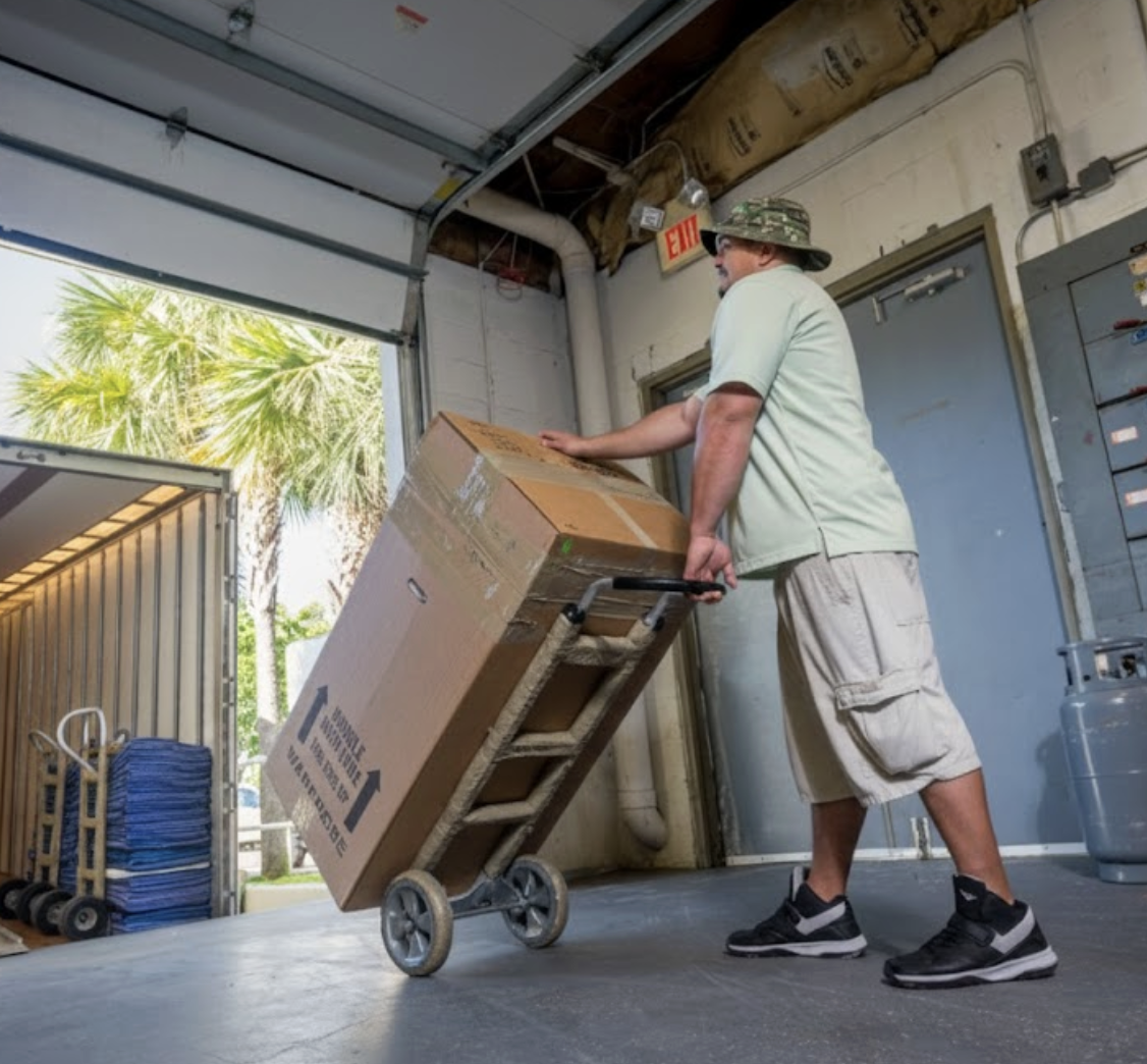 Professional mover using a hand truck to transport a large wardrobe box from a warehouse into a moving truck