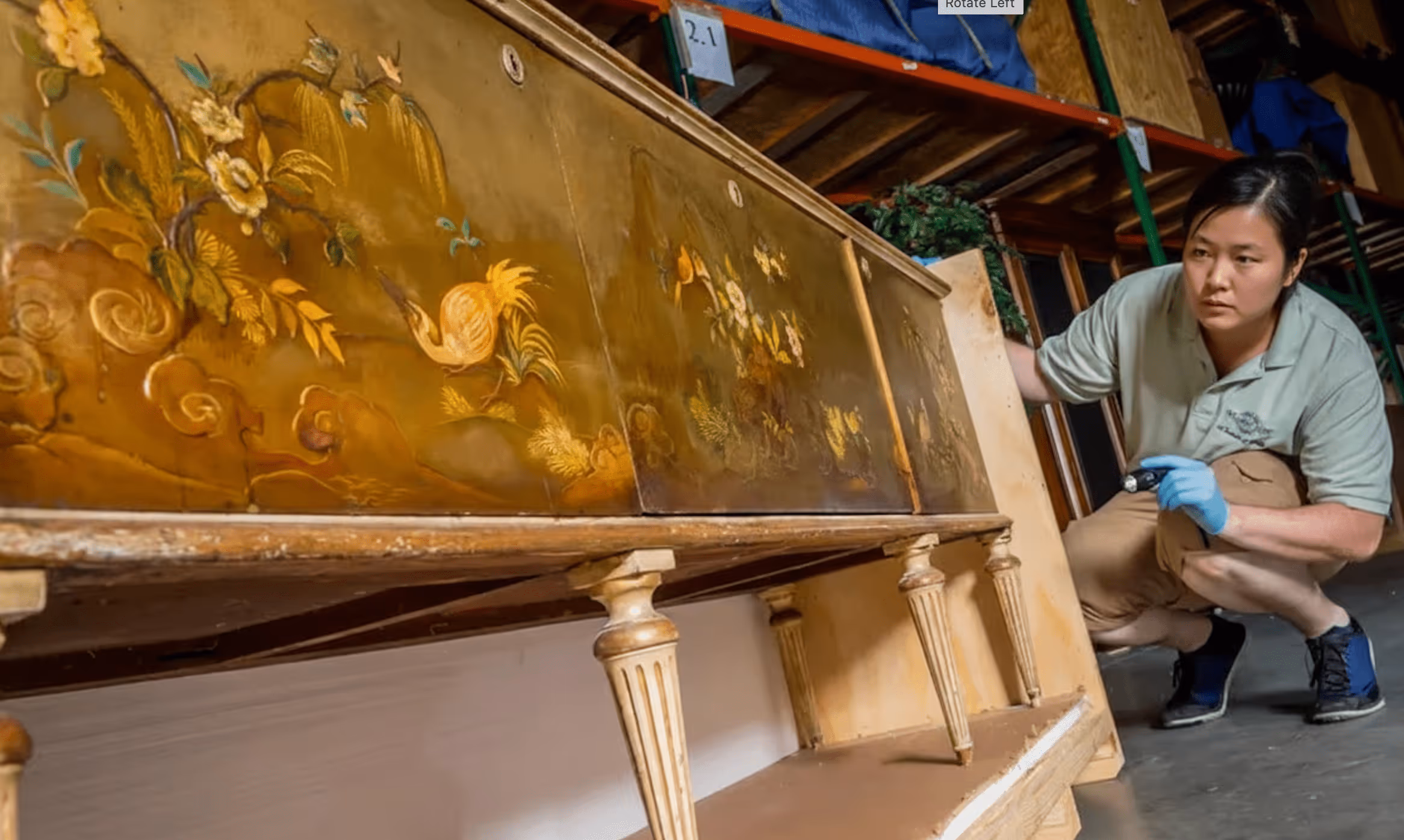 A warehouse specialist wearing gloves inspecting an antique hand-painted sideboard with a flashlight.