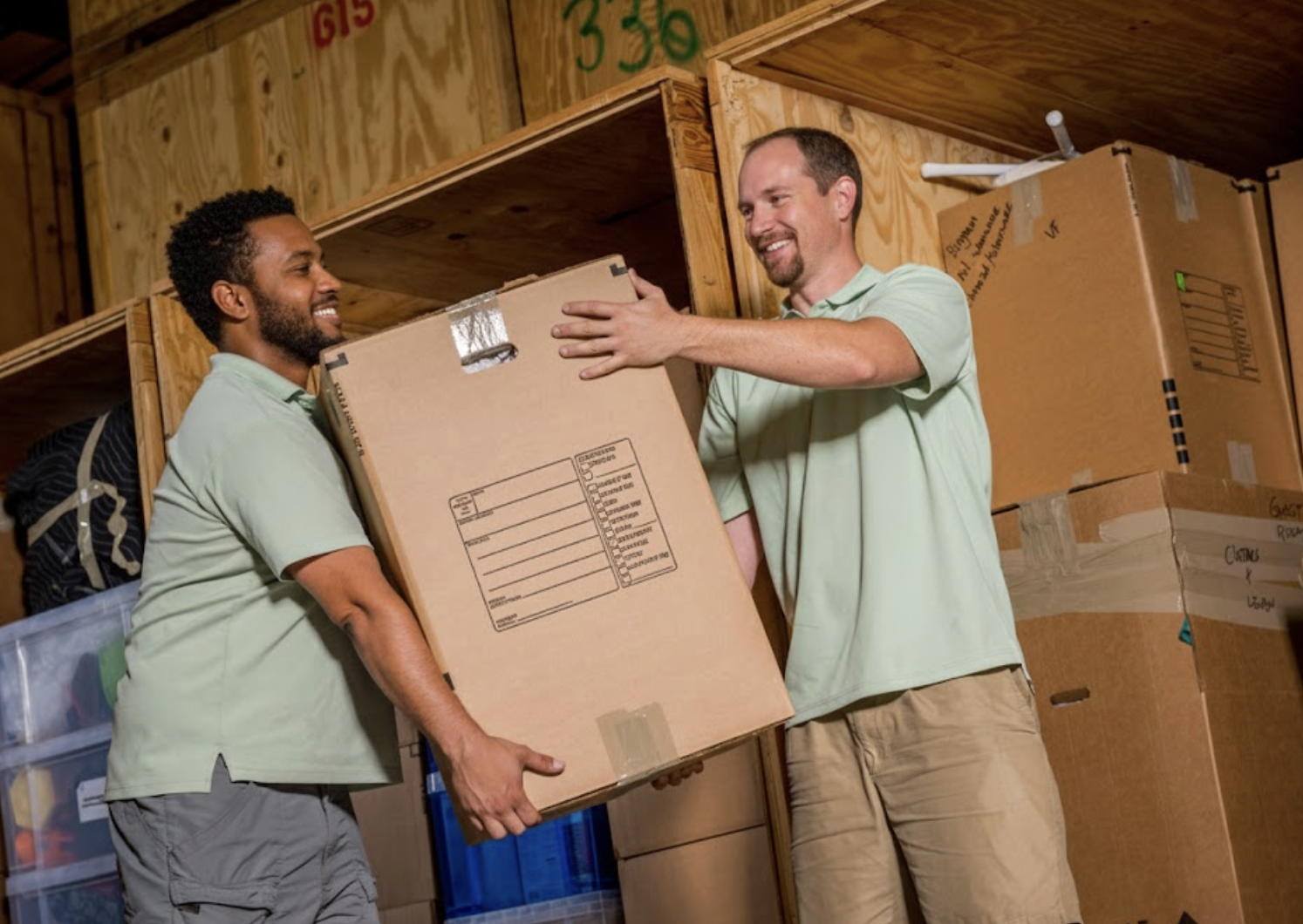 Two professional movers smiling while loading a cardboard box into a wooden storage vault