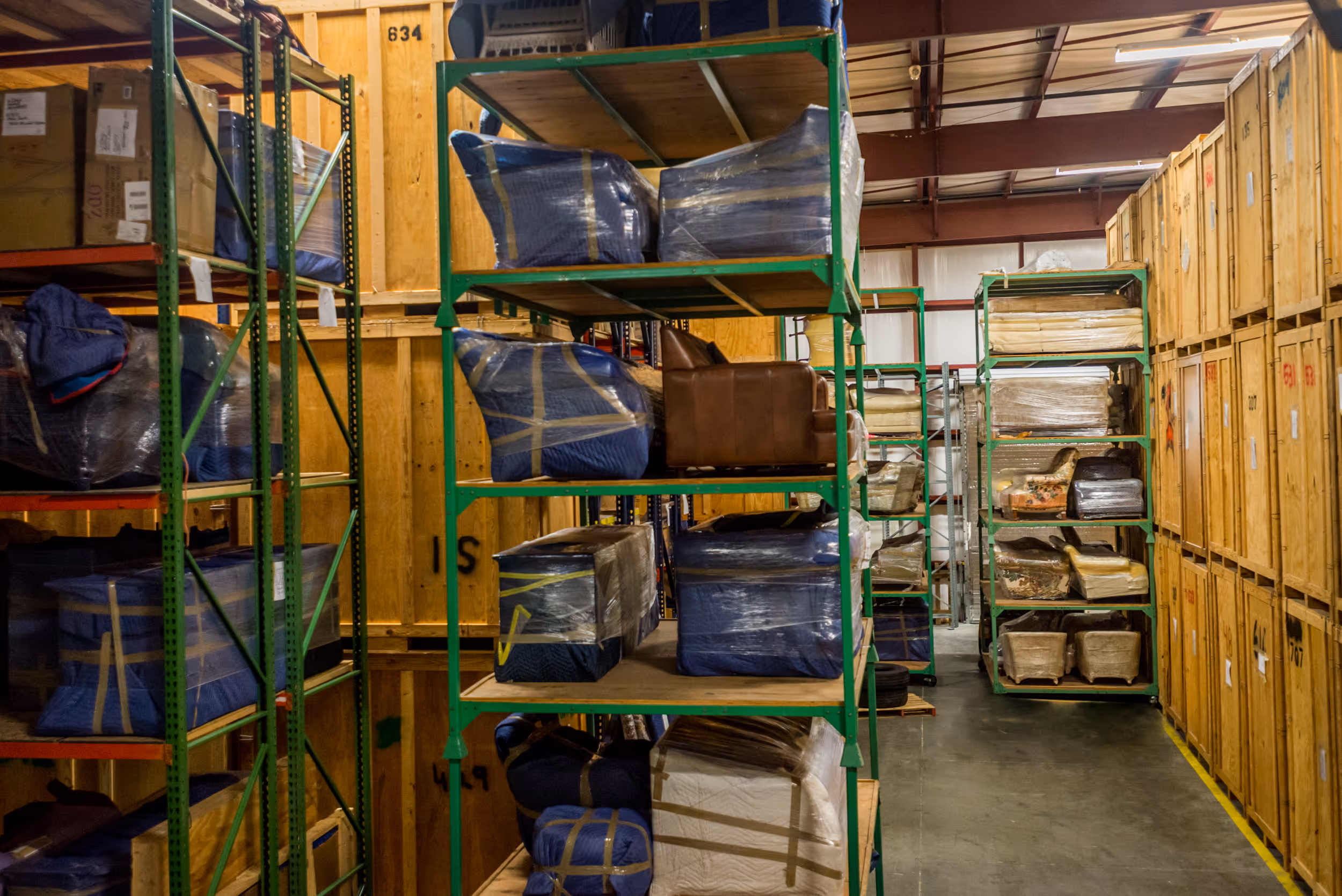 A warehouse storage facility with furniture wrapped in protective blankets on metal shelving beside wooden crates