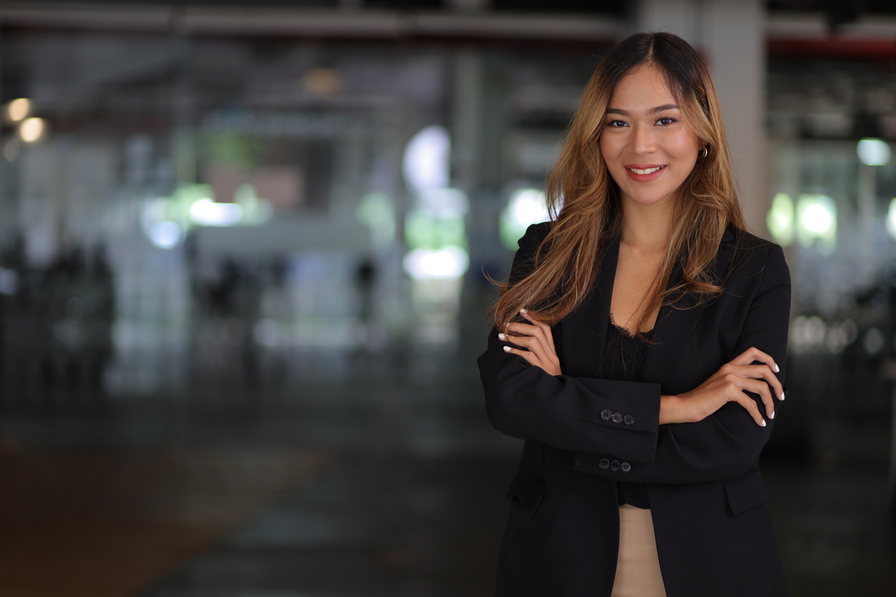 A confident young professional woman in a black blazer standing with arms crossed in a modern office setting