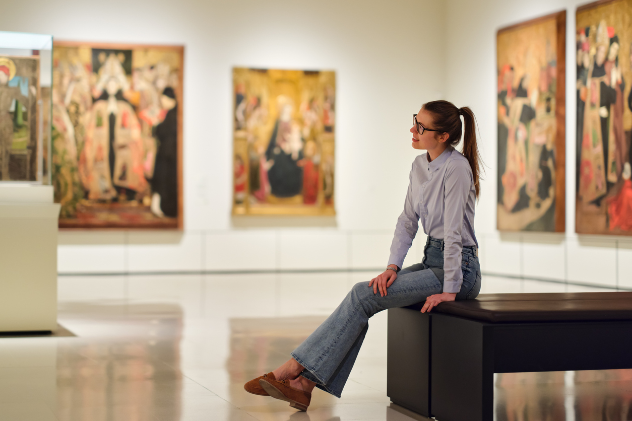 A young woman sitting on a gallery bench viewing classical paintings in an art museum