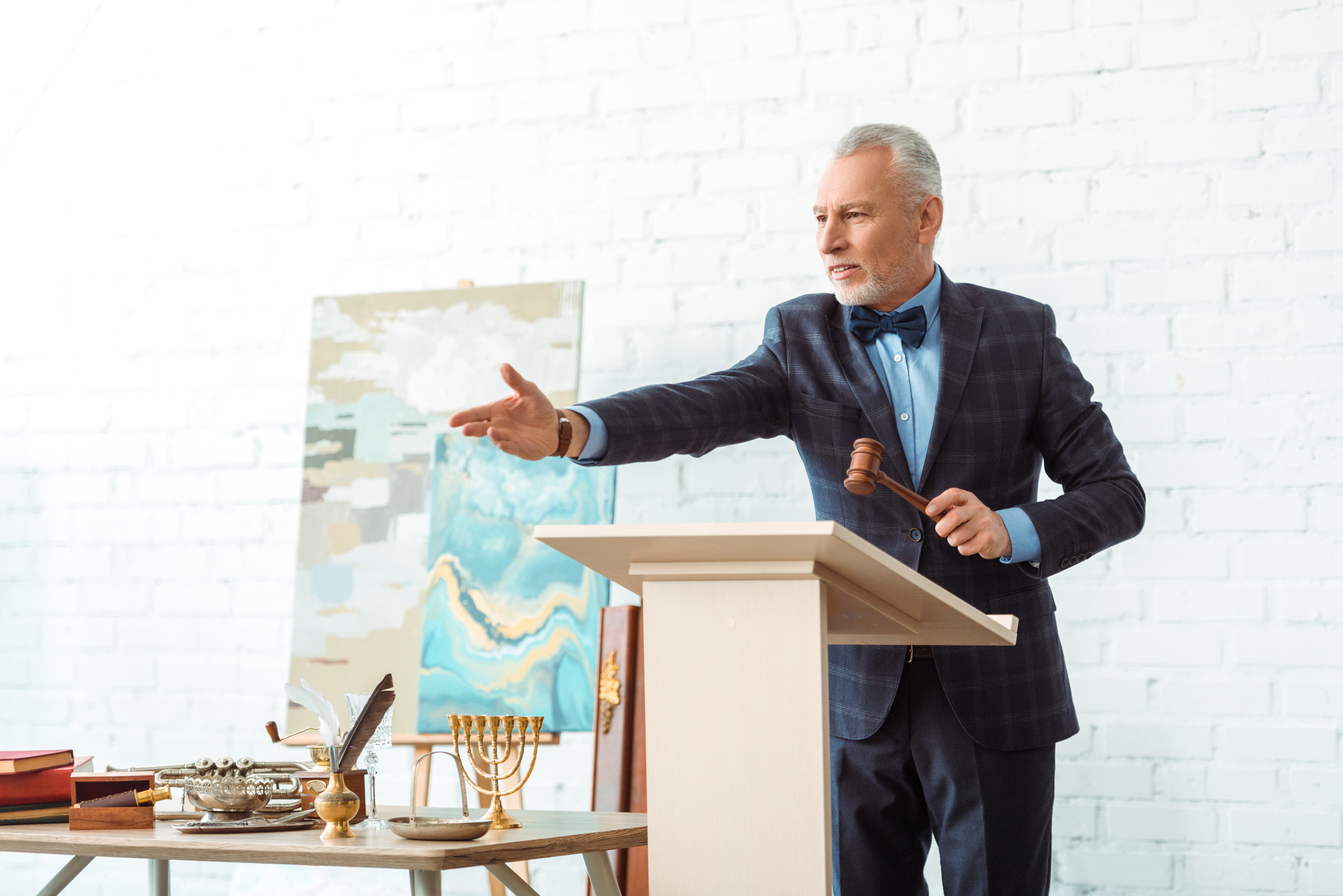 Professional auctioneer in a suit holding a gavel and gesturing at a podium during an art and antiques auction