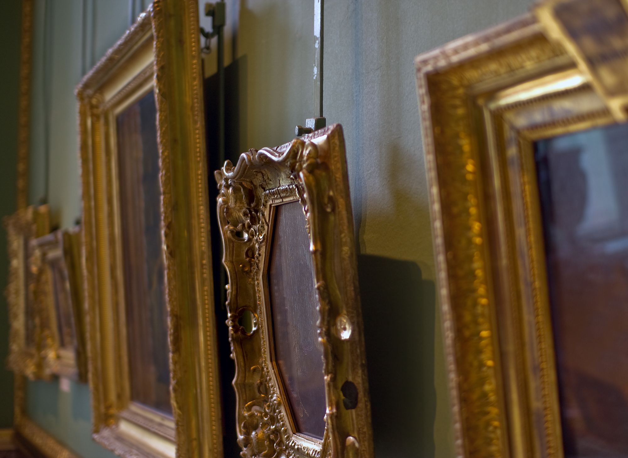 Close-up side view of ornate gold-framed paintings hanging on a gallery wall using a traditional rod-and-rail picture hanging system