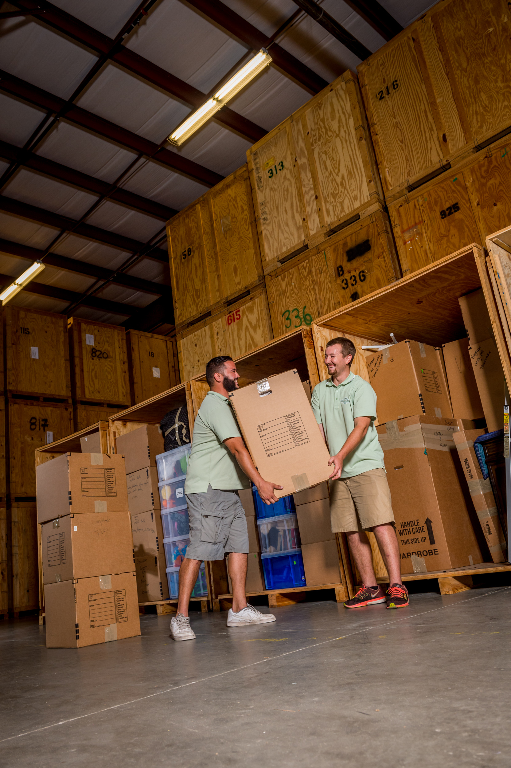Two professional movers loading a large box into a wooden storage vault inside a secure warehouse