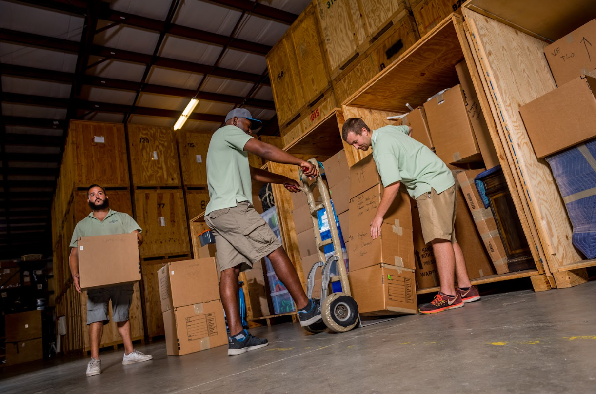 Professional movers loading cardboard boxes onto a hand truck in a storage warehouse.